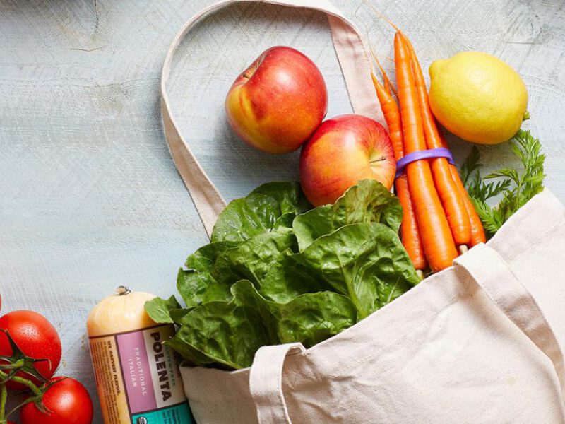 A canvas tote bag full of fresh produce lies on a white counter with more veggies surrounding it