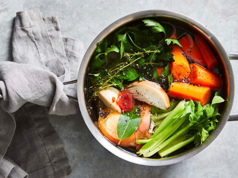 Vegetable Broth full of carrot chunks, celery, an onion wedge, and fresh herbs in a medium silver pot on a gray table with a gray cloth wrapped around the pot handle