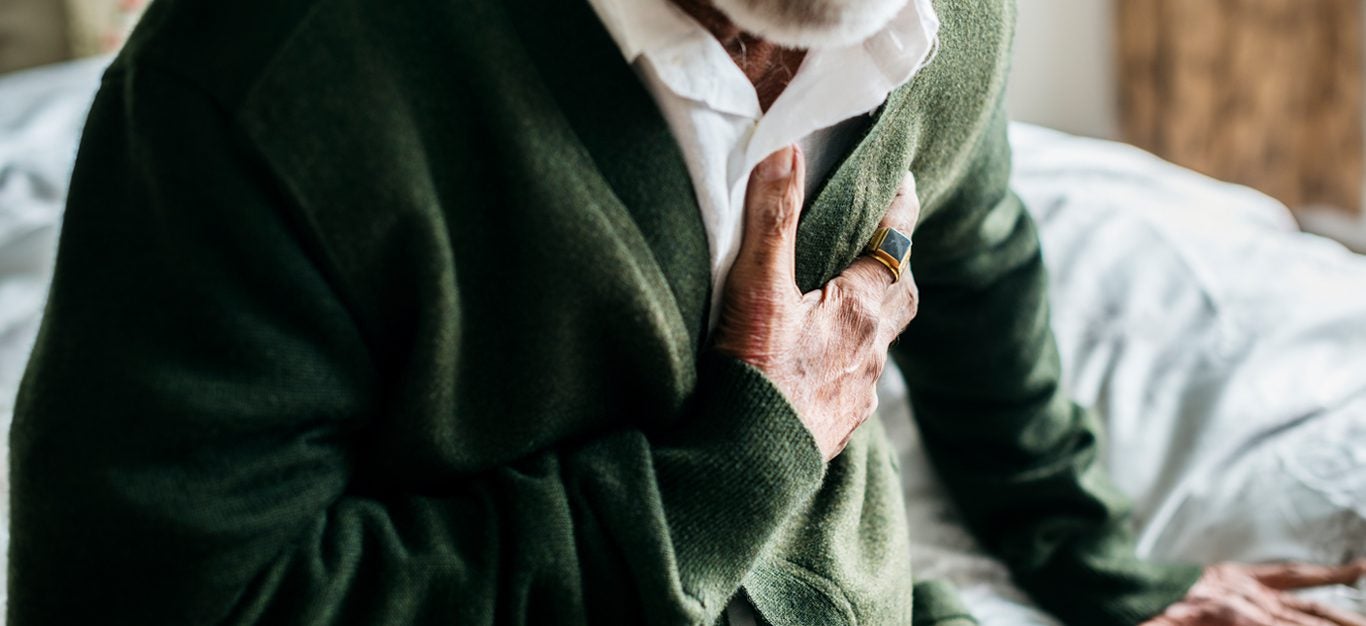 elderly man wearing green cardigan and sitting on bed having chest pains, face not visible