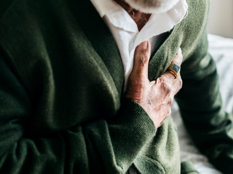 elderly man wearing green cardigan and sitting on bed having chest pains, face not visible