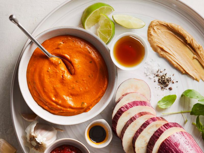 Light Thai Peanut Dressing in a white bowl surrounded by eggplant, soy sauce, peanut butter, and lime slices