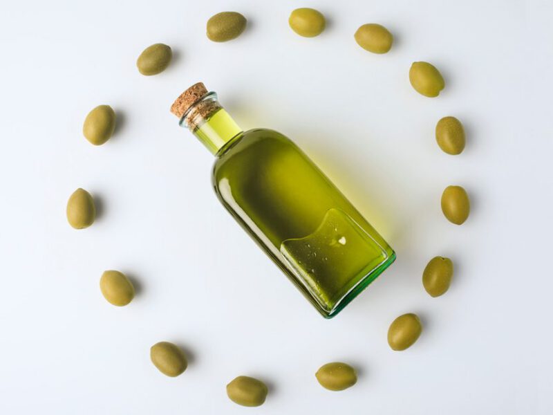 A corked clear glass bottle of olive oil lies on its side surrounded by a circle of green olives