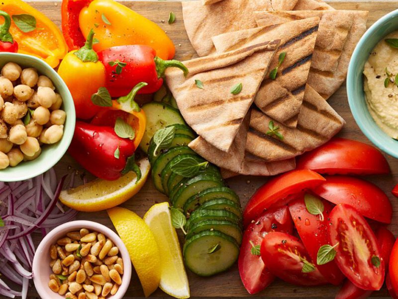 greek salad board with chopped veggies, pita bread, and bowls of hummus on a wooden serving board