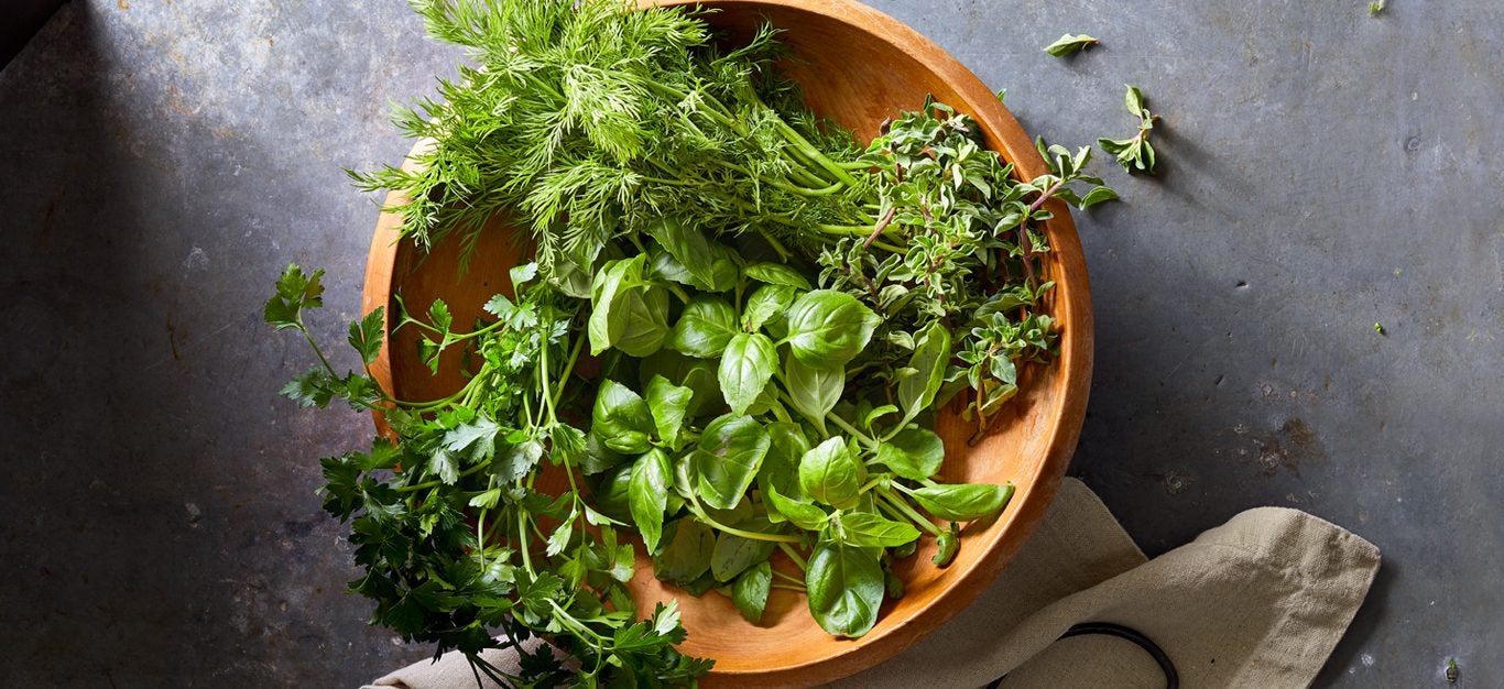 A bowl of snipped fresh green herbs, including mint, basil, oregano, and chives, sitting in a terra cotta bowl on a gray table