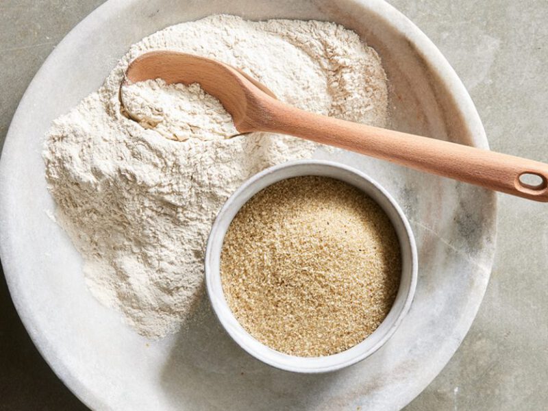 A flat-lay shot of a plate with some ground fonio flour on it, a wooden spoon, and a small bowl with fonio grains in it