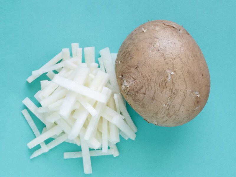 A whole bulb of the root vegetable jicama, on a light blue background, with matchstick-cut slices of peeled jicama beside it