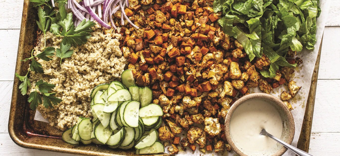 A baking sheet with a colorful array of different ingredients for a vegan shawarma bowl, including roasted sweet potato, diced cucumber, short grains, chopped lettuce, and a small ramekin of tahini sauce on the side