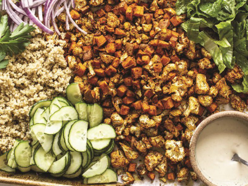 A baking sheet with a colorful array of different ingredients for a vegan shawarma bowl, including roasted sweet potato, diced cucumber, short grains, chopped lettuce, and a small ramekin of tahini sauce on the side
