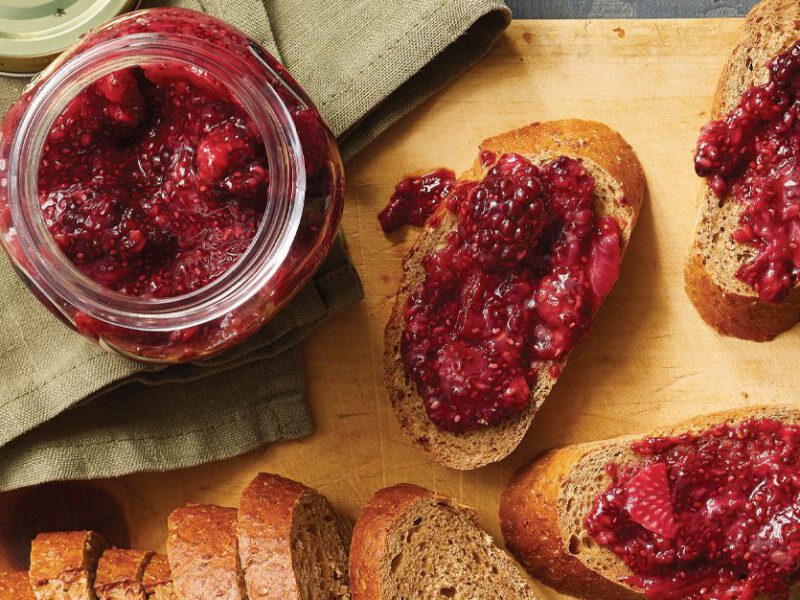 Homemade mixed blackberry, raspberry, and strawberry jam spread on a few slices of whole grain bread with a jar of it off to the side