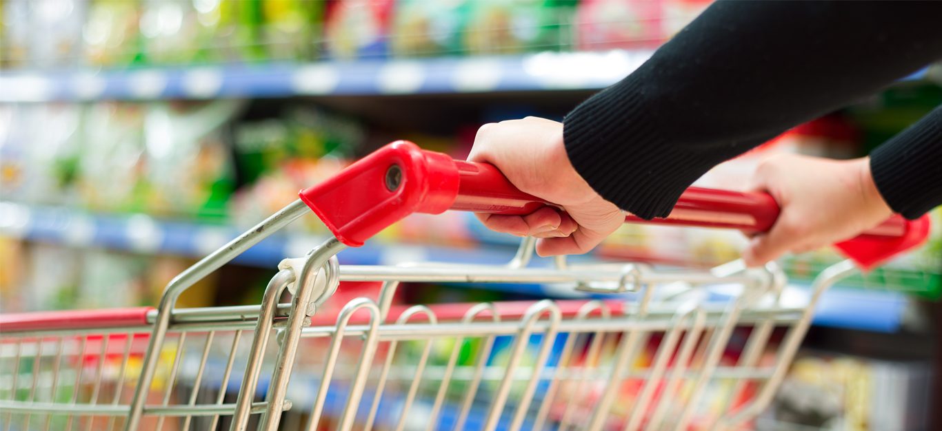 Hands on the red handle of a shopping cart, blurred grocery aisle in the background