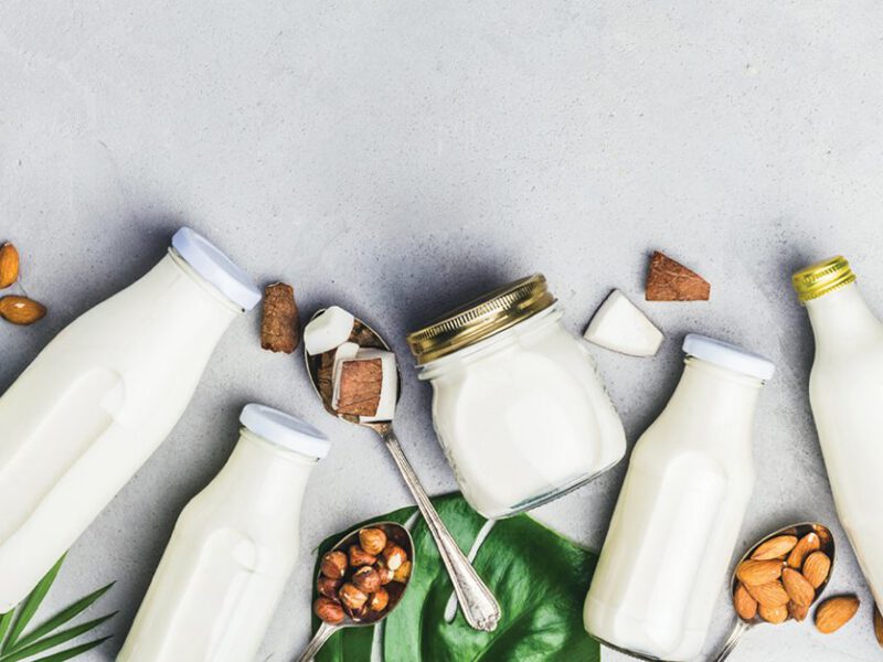 Glass bottles of various types of nondairy plant milks, including oat, soy, and nut, shown laid next to each other, with nuts, soybeans, and leaves in between