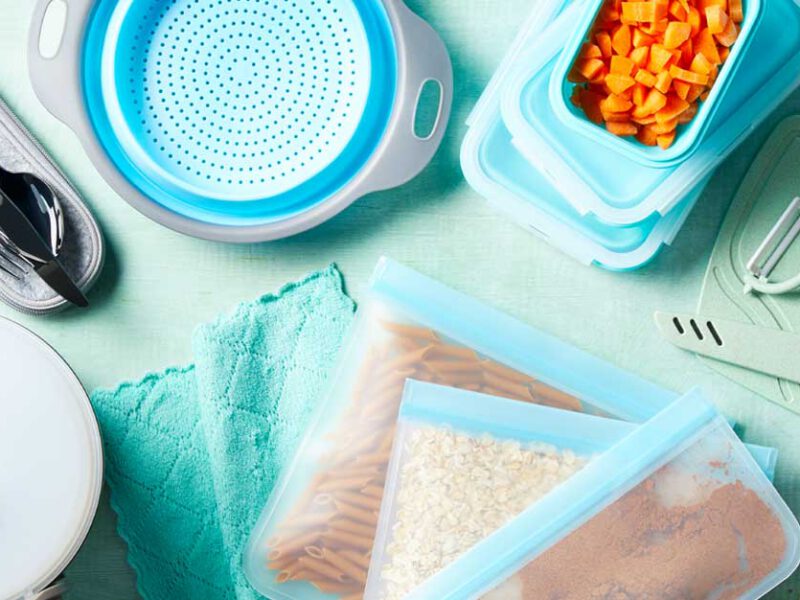 Utensils, resusable bag filled with grains and pasta, a colander, and glass tupperware filled with carrots on light blue tabletop