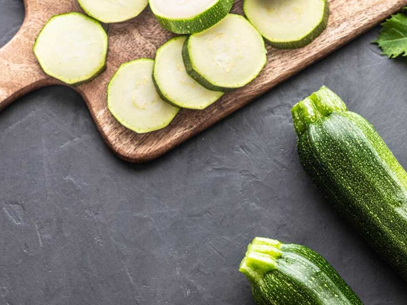 Sliced zucchini on a cutting board with whole zucchini and squash blossoms off to the side at an angle