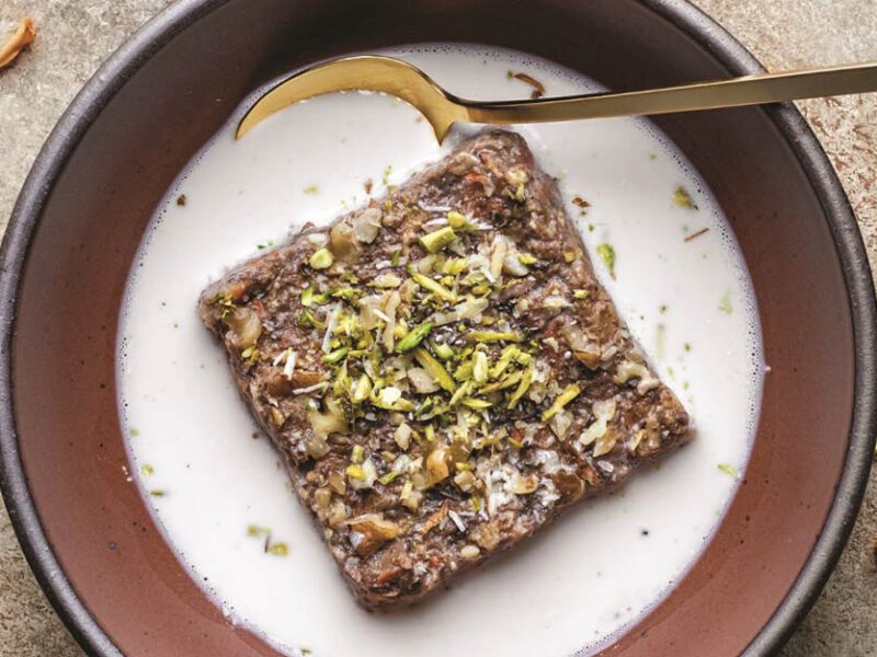 Gajjar No Halvo Baked Oatmeal in a brown bowl, with white milk surrounding the oatmeal square; a garnish of flower petals is visible around outside of the bowl