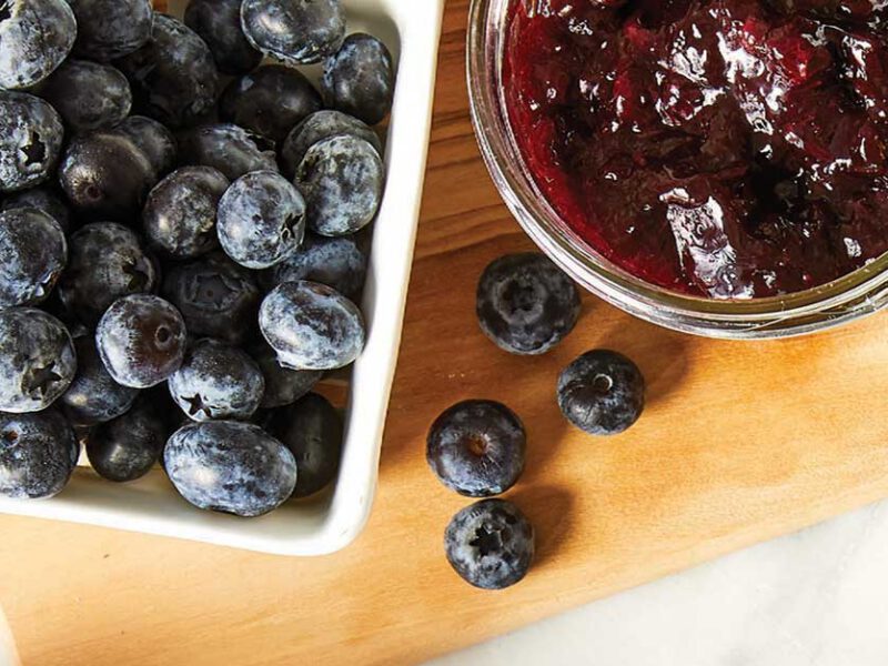 A small carton of blueberries beside a mason jar of homemade jam on a cutting board