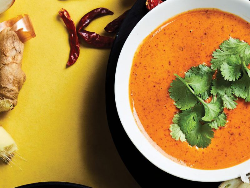 Thai Red Curry Sauce in a white bowl against a yellow background surrounded by lime slices, ginger root, and dried chili peppers