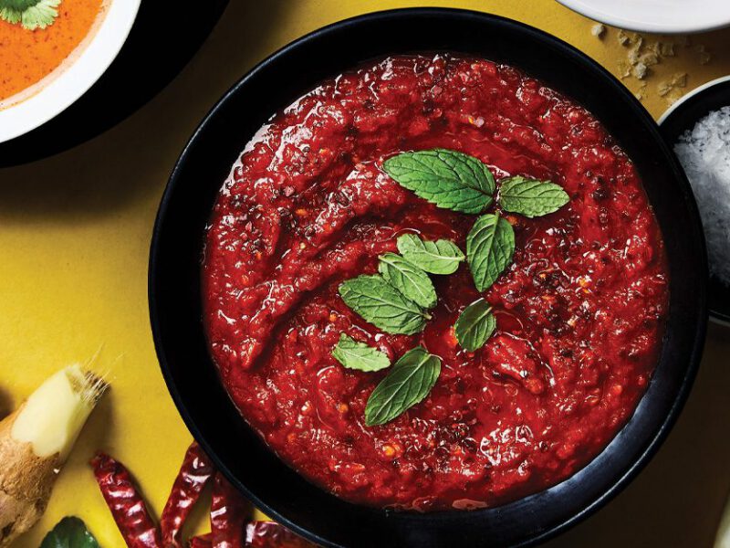 Homemade Harissa in a dark bowl against a yellow background surrounded by fresh ginger, chiles, and onion slices
