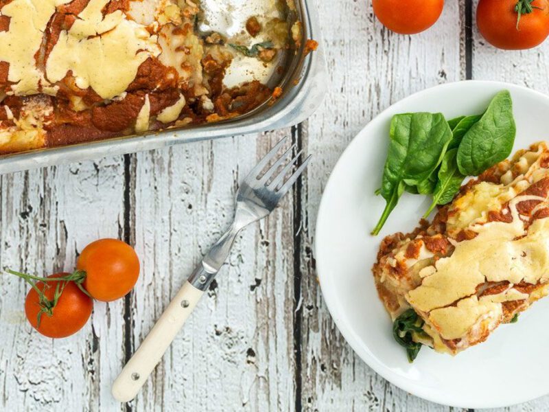 Sweet Potato Lasagna slice on a white plate next to a large tray of lasagna on a white wooden tabletop