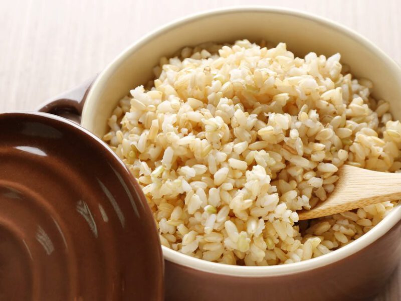 Brown ceramic saucepan full of cooked brown rice, with a wooden spoon nestled beneath the rice