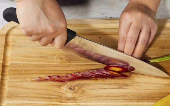 Hands hold a knife cutting a purple carrot into a bias slice atop a wooden cutting board