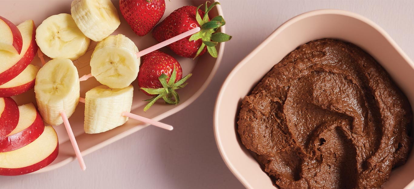 Healthy Chocolate Hummus in a light pink bowl next to a plate of fresh fruit slices