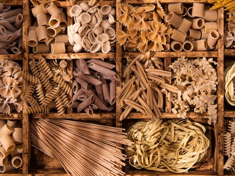 An array of whole wheat pasta shapes in a wooden crate, with compartments dividing each shape, including penne, orecchiette, spaghetti, and rotini