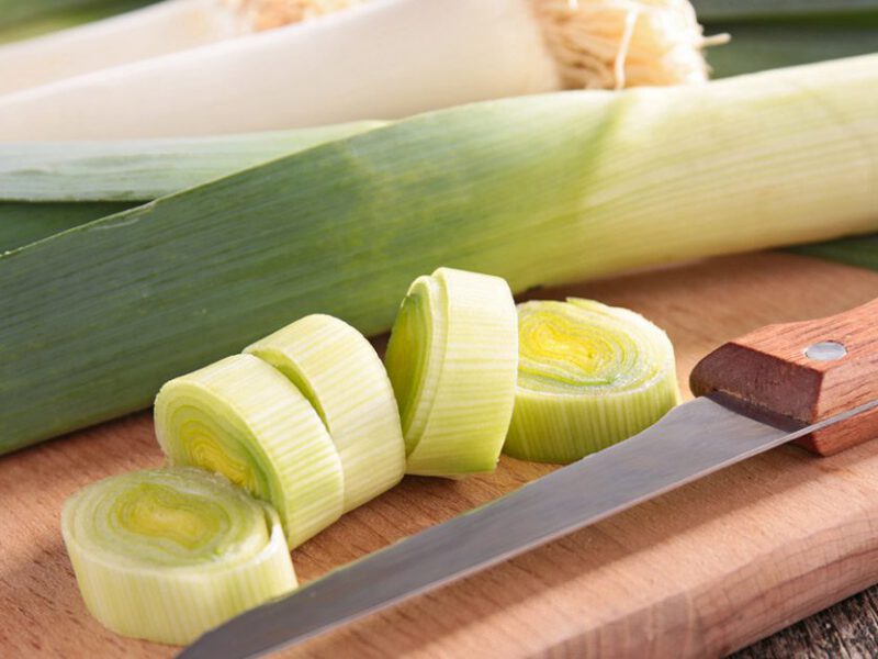 close up shot of sliced leeks on a cutting board beside whole leeks, with a knife to the side