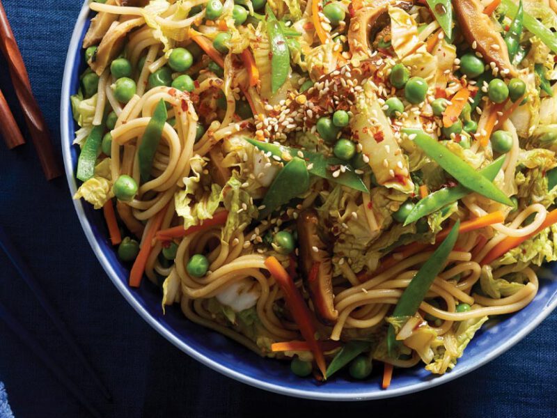 Stir-Fried Udon Noodles with Mixed Vegetables in a blue bowl with chopsticks and small bowl of red pepper flakes to the right