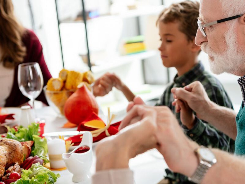 Multiple generations of a family gathered around a not plant-based holiday meal saying grace
