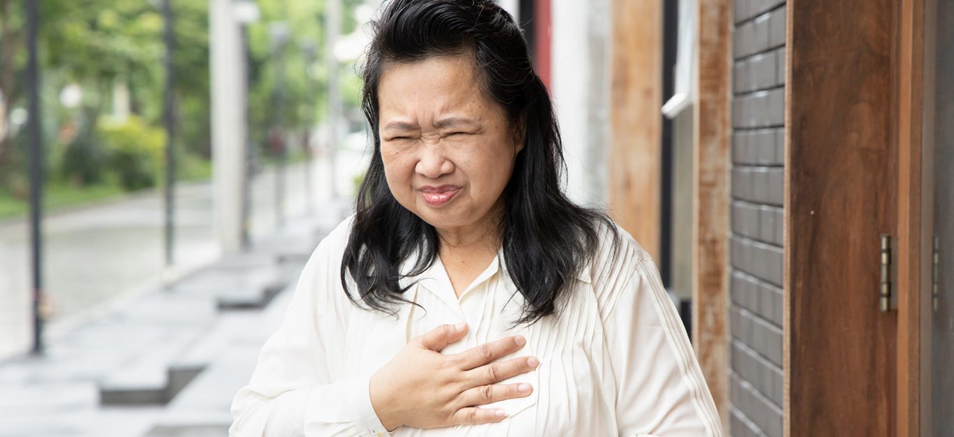 Asian woman experiencing GERD, chronic acid reflux, standing on a sidewalk and holding her chest