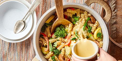A pot of pasta with creamy vegan sauce and Broccolini, with a hand pouring additional sauce over it