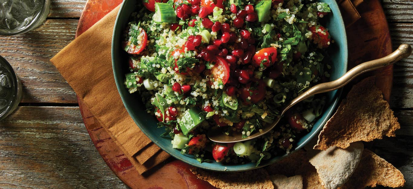Quinoa Tabbouleh with Pomegranate Seeds in a blue dish with a gold metal spoon and a side of crackers