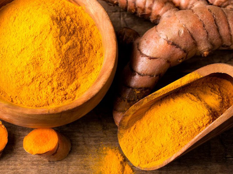 A table with an array of turmeric on it, including a wooden bowl of dried ground turmeric, with a scoop next to it filled with more dried turmeric, and a knob of fresh turmeric root and some fresh turmeric slices