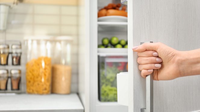Woman's hand opening the door of a fridge to reveal produce with bins of dry goods on the counter in the background