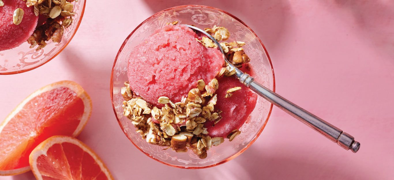 Grapefruit Nice Cream with Skillet Granola in a round glass with a metal spoon against a pink background