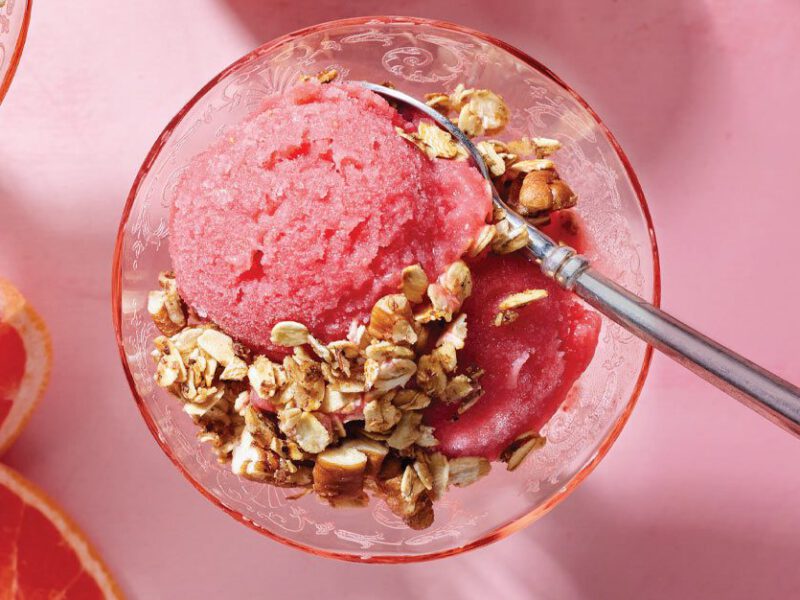 Grapefruit Nice Cream with Skillet Granola in a round glass with a metal spoon against a pink background