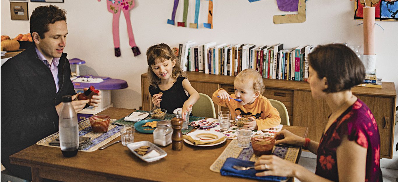 A man and woman sit at opposite ends of a table, with two young children sitting between them, enjoying a plant-based meal as a family