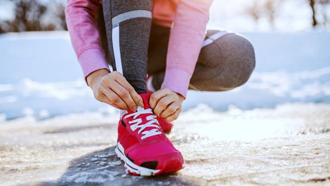 woman in a pink top and red sneakers ties her shoe on a snowy road