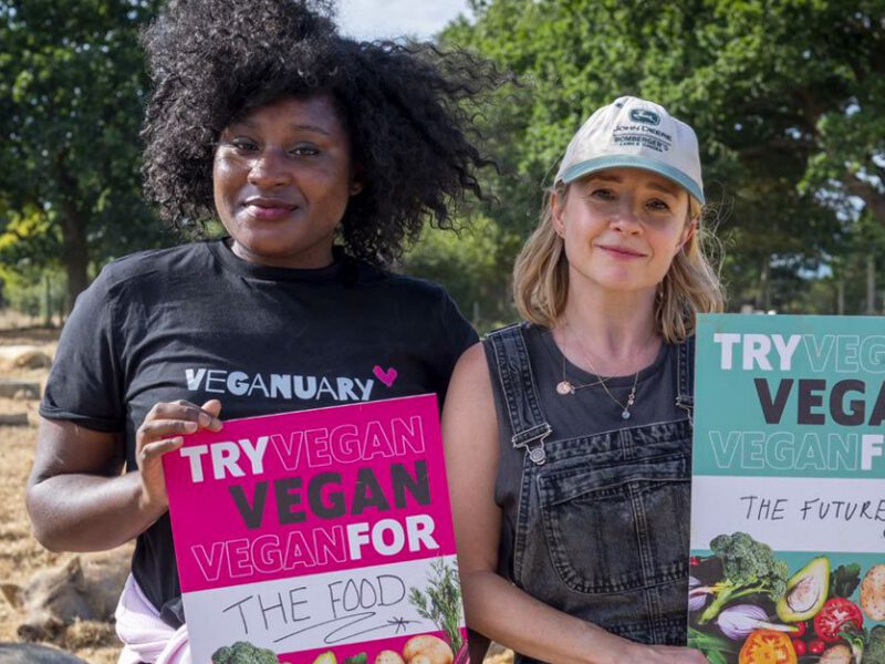 Two women standing in an animal sanctuary holding signs that say Try vegan for the food, and Try vegan for the future