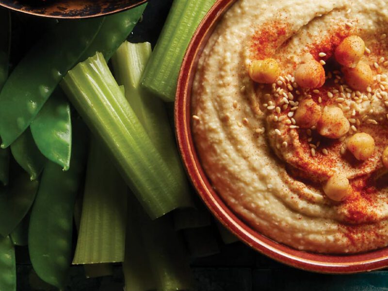 Low-Fat Hummus in a red bowl next to celery sticks and snap peas