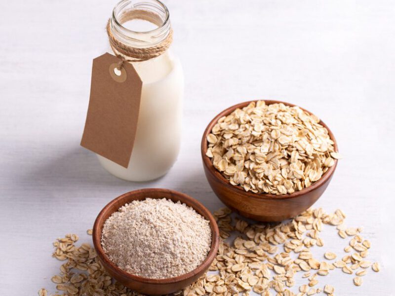 A clean white surface with a carafe of oat milk, a small bowl of oat flour, and a slightly larger bowl of whole oats