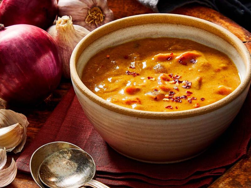 Shiro Wat (Chickpea Flour Stew) in a white ceramic bowl surrounded by red onions and garlic on a wood tabletop