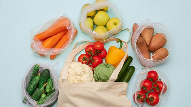 A bag of fresh vegetables spilling out on a flat blue surface, surrounded by five smaller bags, containing bags of cucumber, carrot, apples, potatoes, and tomatoes