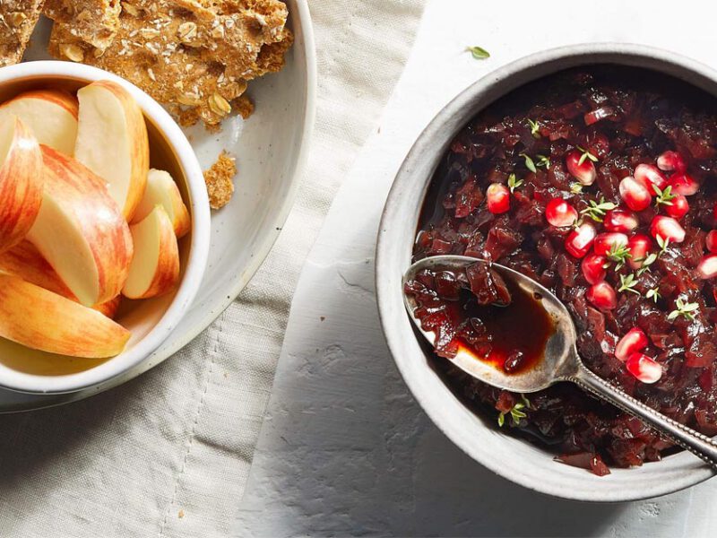 Red Onion Confit in a small gray bowl with a metal spoon next to a plate of crackers and apple slices