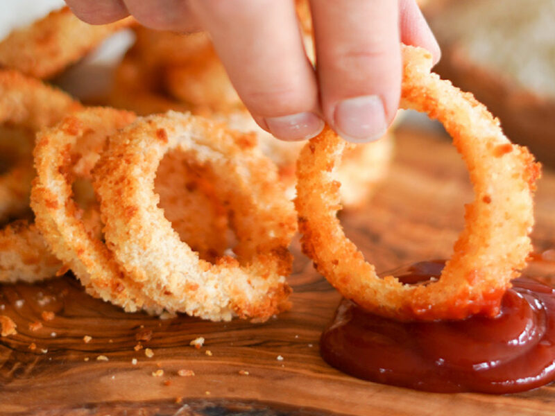 A photo of baked or air-fryer onion rings on a wooden tray, with a hand holding one ring and dipping it in ketchup