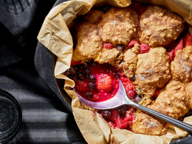 Bursting with Berries Cobbler in a cast iron skillet lined with parchment paper with a big metal spoon