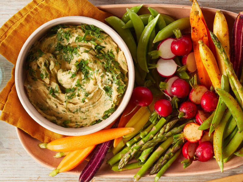 Two-Way Vegetable Platter with Herbed Hummus in a tan ceramic dish with yellow cloth napkins next to two blue water glasses