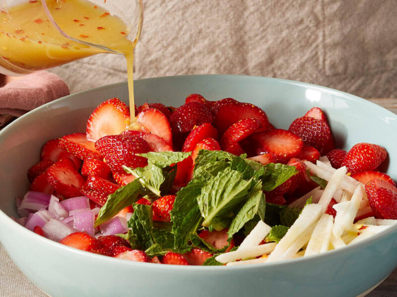 Strawberry-Mint Salad with Easy Maple-Dijon Dressing in a light blue ceramic bowl with a woman's hand holding the right side of the bowl