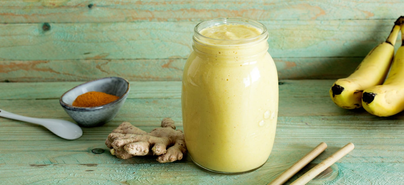 Pineapple Ginger Smoothie in a glass jar next to fresh ginger root and turmeric against a green wood backdrop