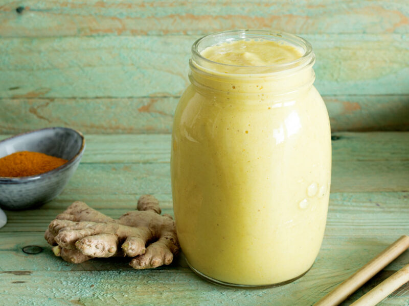 Pineapple Ginger Smoothie in a glass jar next to fresh ginger root and turmeric against a green wood backdrop